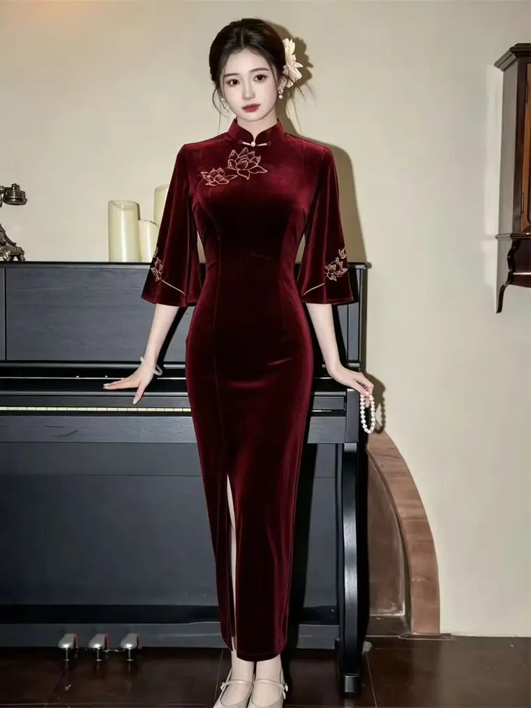 A woman wearing a long, form-fitting velvet cheongsam-style dress in deep burgundy with floral embroidery near the neckline and sleeve cuffs, standing indoors beside a black piano.