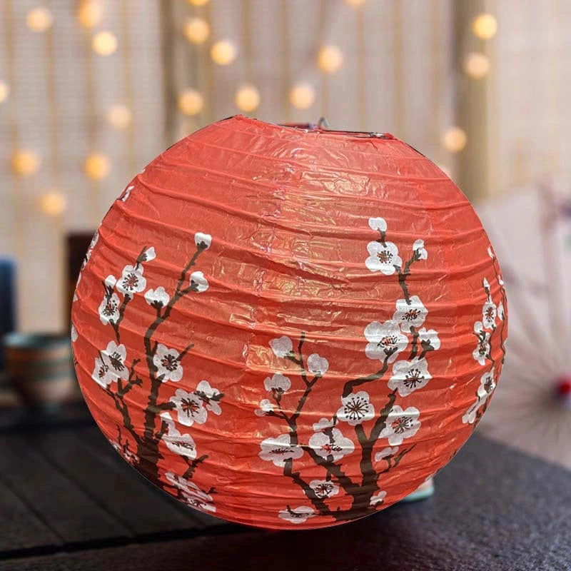 A round red Chinese paper lantern decorated with white cherry blossoms and dark brown branches, resting on a dark surface with a softly blurred background of warm string lights.