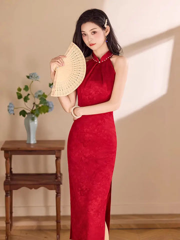 A woman wearing a red Pearl-Trimmed Sleeveless Halter Jacquard Satin Qipao with a high neck and side slit, holding a beige woven folding fan in a sunlit indoor setting.
