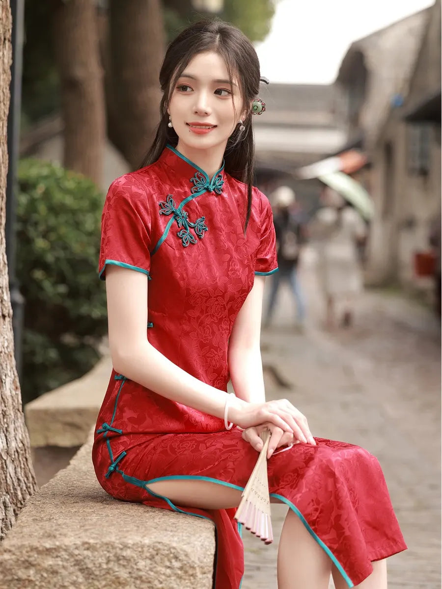 Woman in a red traditional Short Sleeves Floral Jacquard Long Qipao Dress With Blue Trim sitting on a stone bench outdoors.