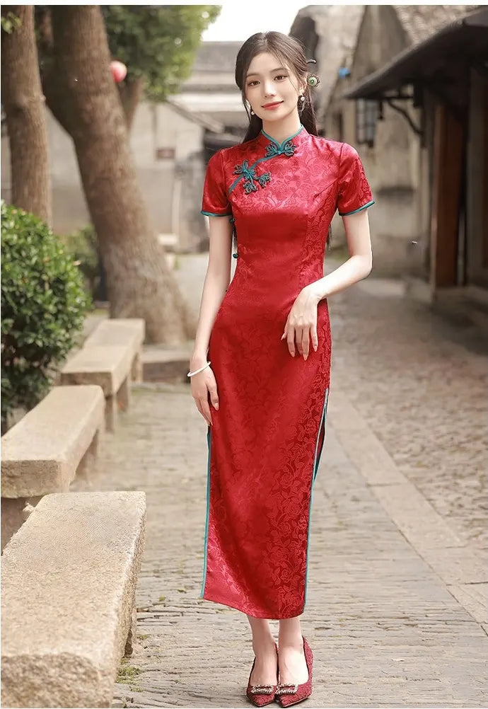 Woman in a red traditional Short Sleeves Floral Jacquard Long cheongsam Dress With Blue Trim standing on a stone path with greenery and a building in the background.