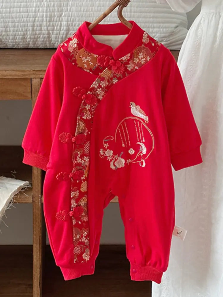 A baby's red long-sleeve romper with traditional Chinese embroidery and decorative buttons, hanging on a wooden hanger against a white curtain and wooden background.