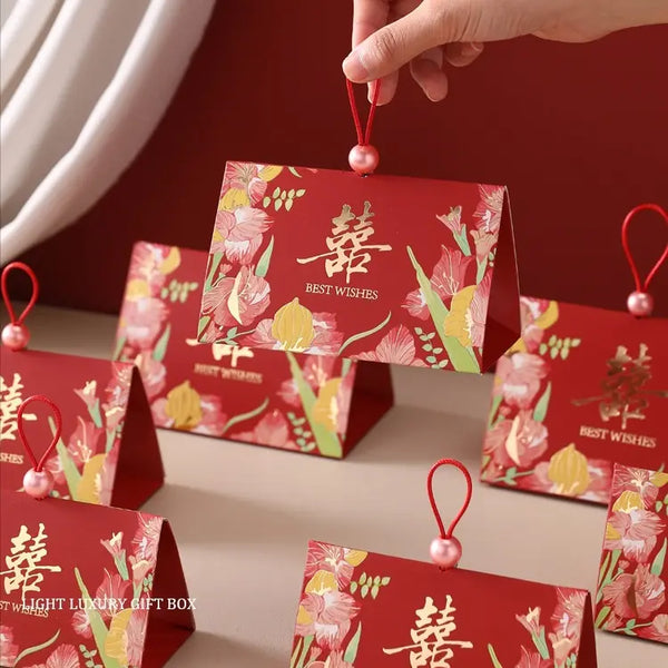 Triangular Chinese  Double Happiness Wedding Favor Boxes with Pearl Loop with floral illustrations and gold Chinese characters, held by a hand against a warm red and beige background.