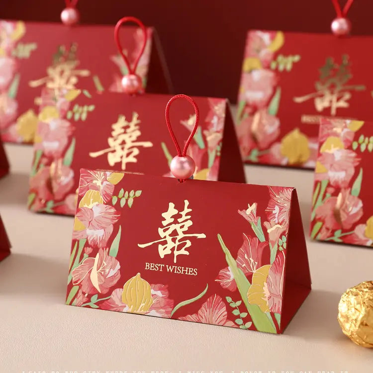 Triangular Chinese  Double Happiness Wedding Favor Boxes with Pearl Loop  with floral illustrations and gold Chinese characters, each hanging from a red loop with a pink bead, arranged on a light surface.