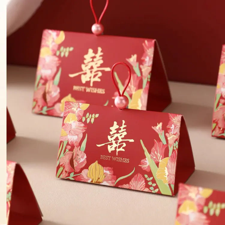 Triangular Chinese  Double Happiness Wedding Favor Boxes with Pearl Loop with floral illustrations and gold Chinese characters, hanging by red strings with pearl beads, shown on a beige surface.