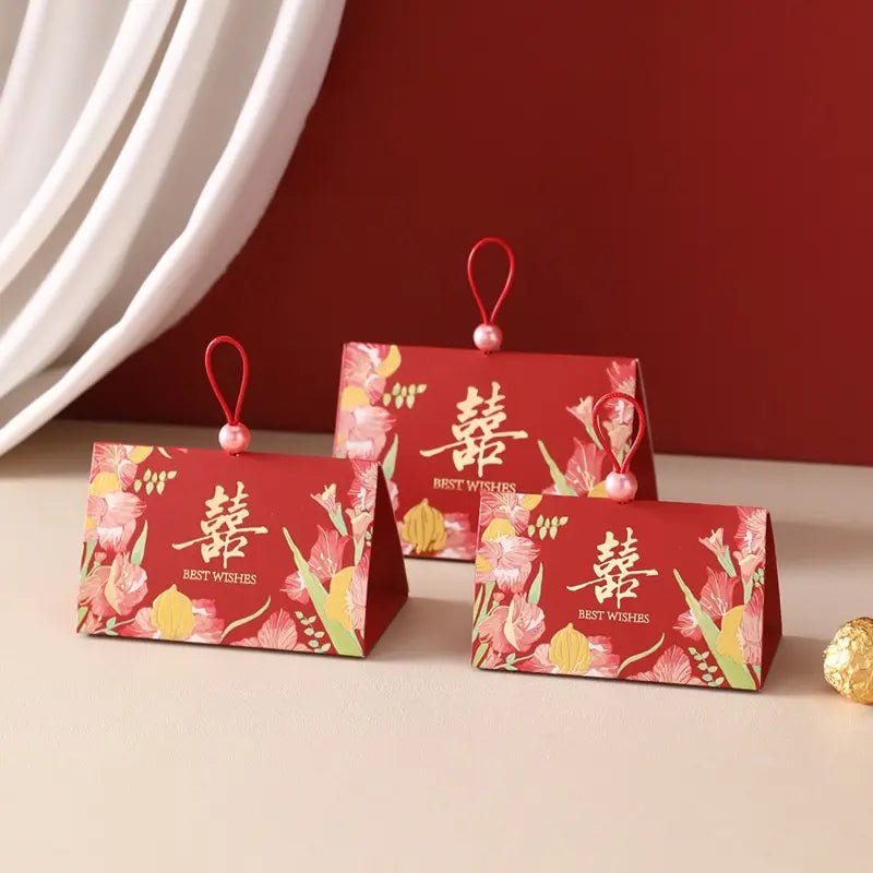 Three red Triangular Chinese  Double Happiness Wedding Favor Boxes with Pearl Loop , decorated with floral illustrations and Chinese characters, arranged on a beige surface against a dark red and white background.
