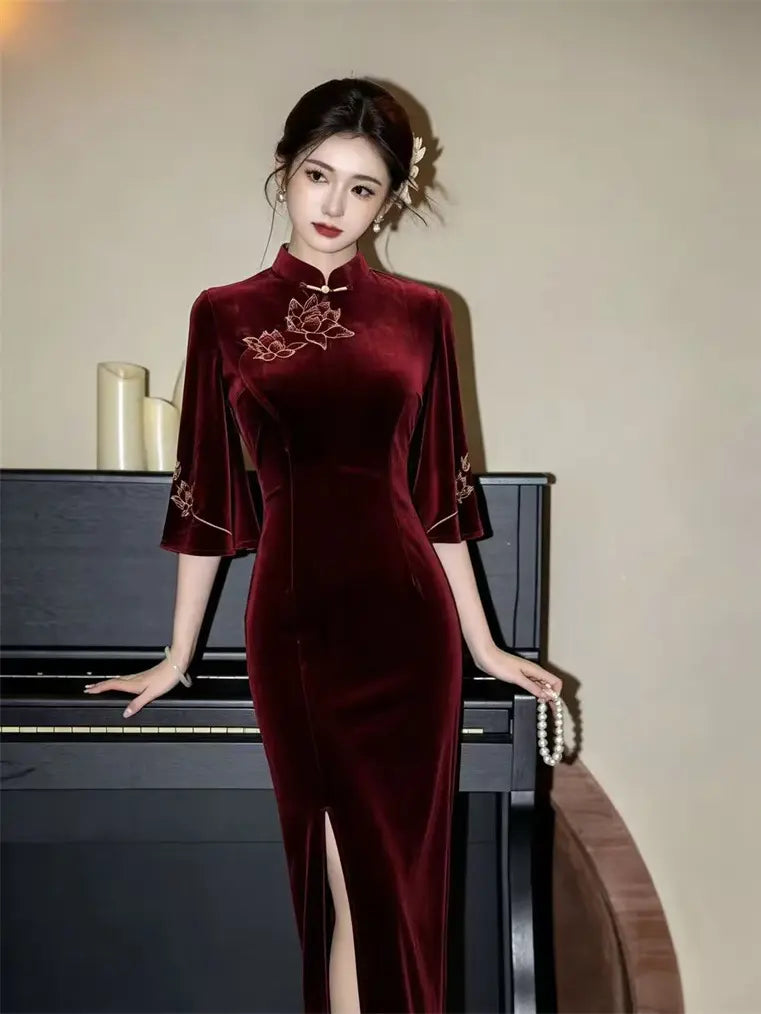 A woman wearing a deep burgundy velvet cheongsam dress with gold floral embroidery on the chest and sleeves, posing in front of a black piano.