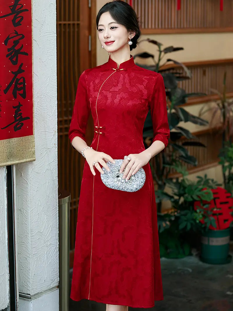 A woman wearing a red a line qipao cheongsam-style dress with three-quarter sleeves and a high collar, holding a silver patterned clutch. She is standing in a decorated indoor setting with Chinese New Year decorations visible in the background.