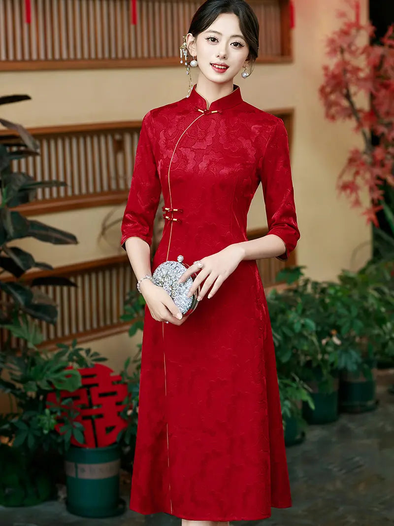 A woman wearing a red Chinese A line cheongsam (qipao) dress with three-quarter sleeves and a high collar, holding a silver patterned clutch purse in a decorated indoor setting with plants.