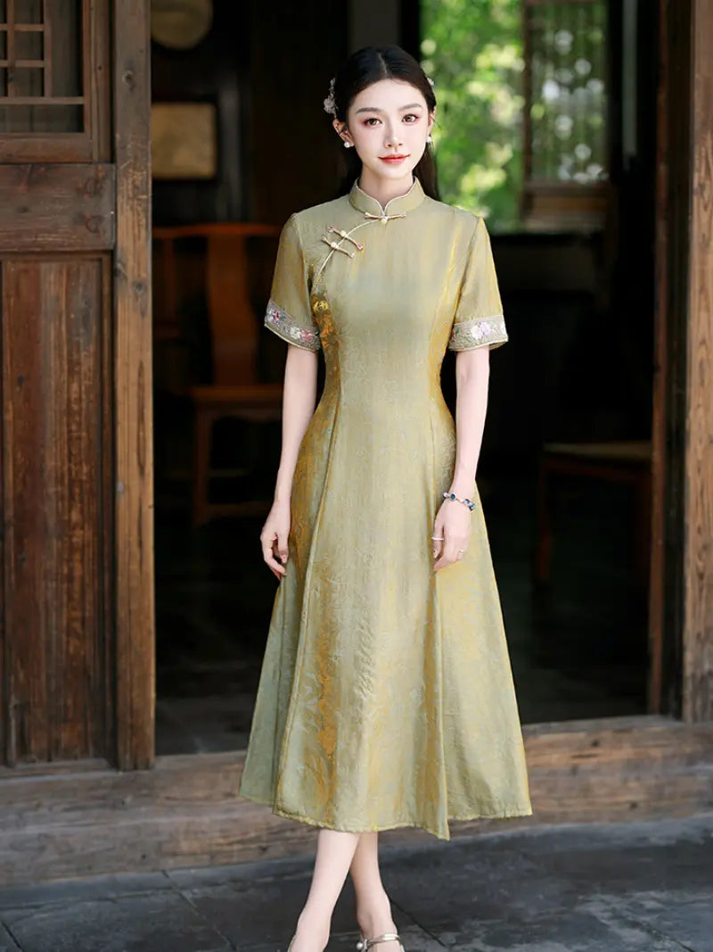 A woman wearing a short-sleeved, midi-length cheongsam-style dress in a muted olive-gold color with gold embroidery on the collar and sleeve cuffs, standing in front of a rustic wooden doorway.