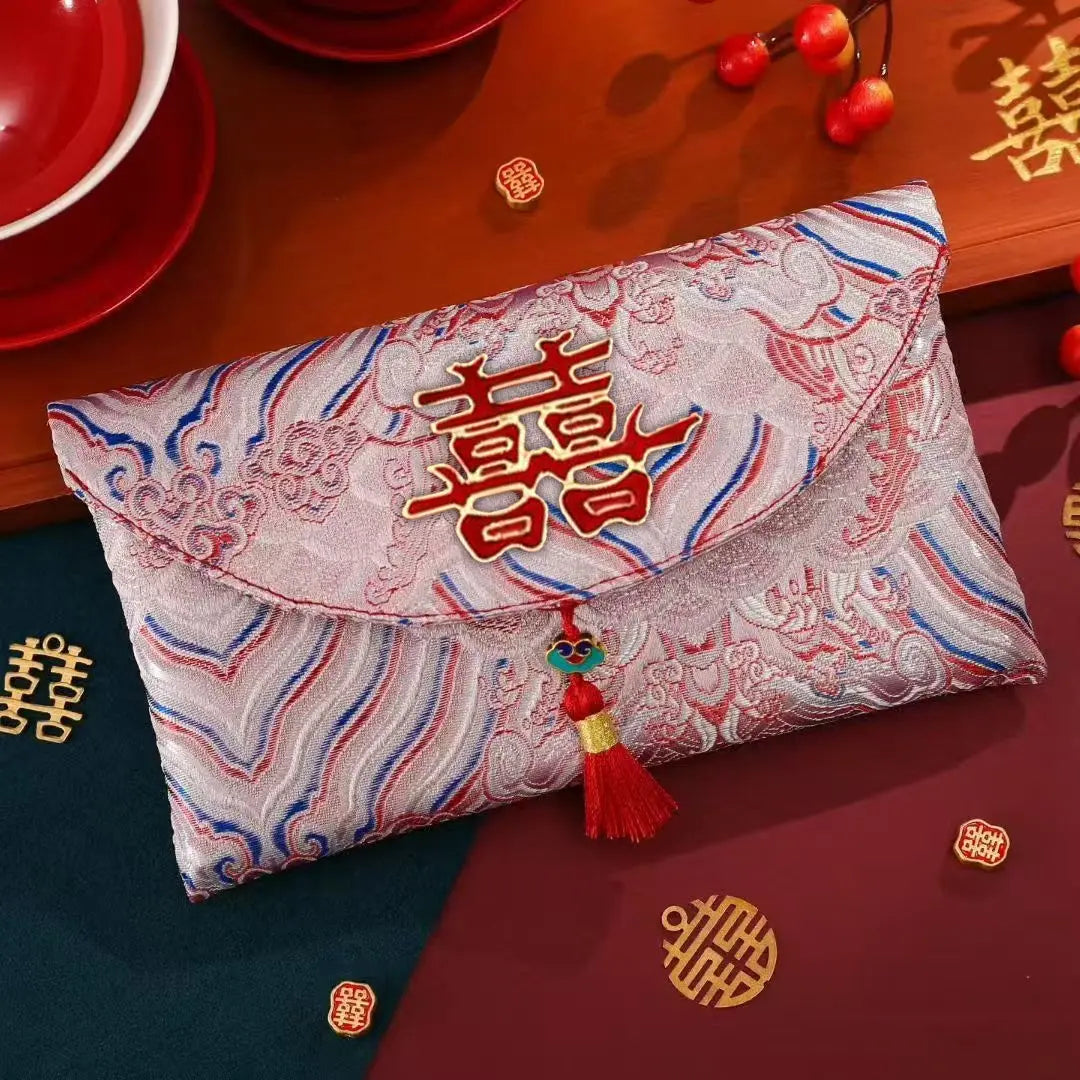 A Chinese wedding envelope clutch purse with a red double happiness symbol on a white brocade patterned fabric, featuring a red tassel charm, displayed on a decorated wooden table.