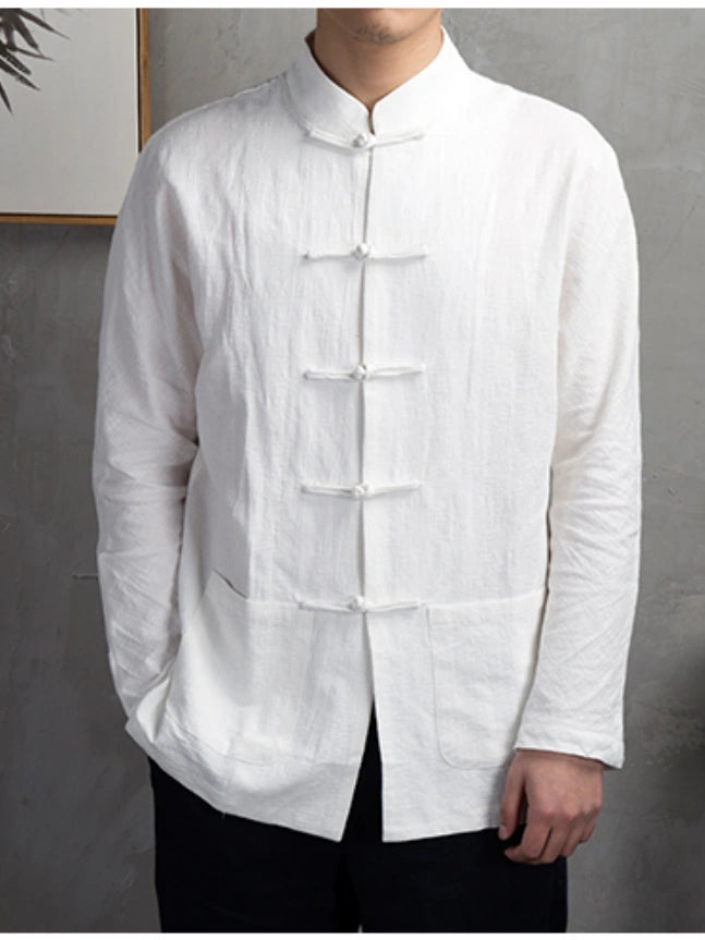 A man wearing a white long-sleeve linen shirt with traditional Chinese frog closures and a mandarin collar, paired with black pants, standing against a gray textured wall.
