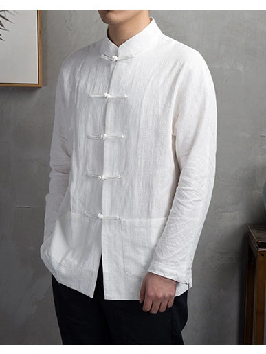 A man wearing a white long-sleeve traditional Chinese-style kongfu shirt with a mandarin collar and buttoned front, paired with black pants, standing against a textured gray wall.