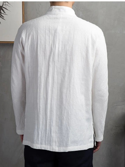 A man viewed from the back wearing a white long-sleeve linen shirt with a high collar, paired with dark pants, set against a textured gray wall.