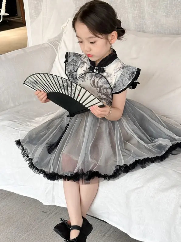 A young girl sits on a white bed wearing a black and gray traditional-inspired Ink Painting Sleeveless Layered Tulle Qipao cheongsam  dress with a sheer tulle skirt and holding a black folding fan.