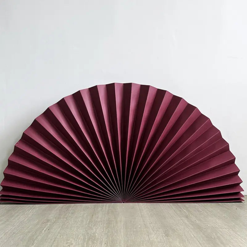 A burgundy pleated folding decorative fan, partially unfolded, resting on a light gray wood floor against a plain white wall.