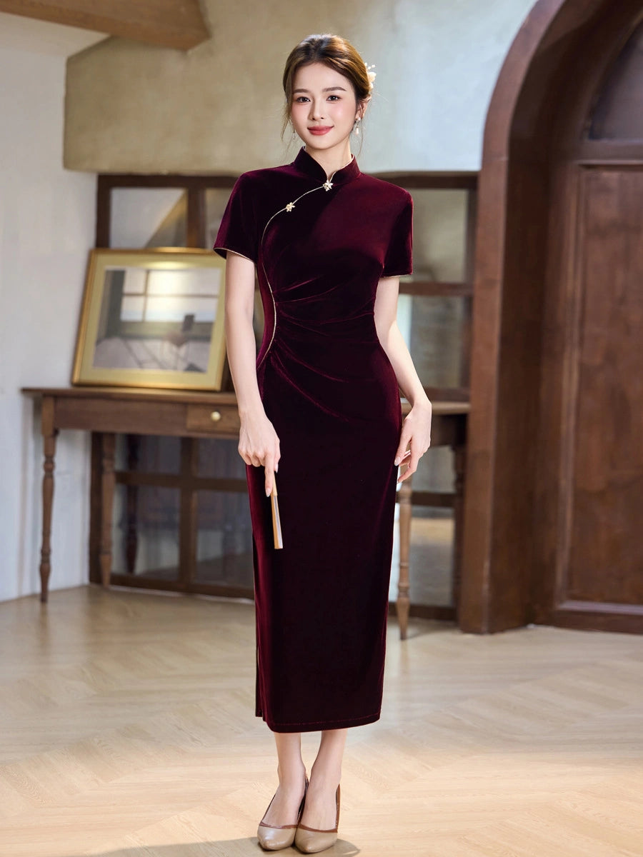 A woman wearing a wine red short-sleeved, high-neck, midi-length velvet cheongsam dress in deep burgundy. She is standing in a warm, rustic interior with wooden floors and furniture.