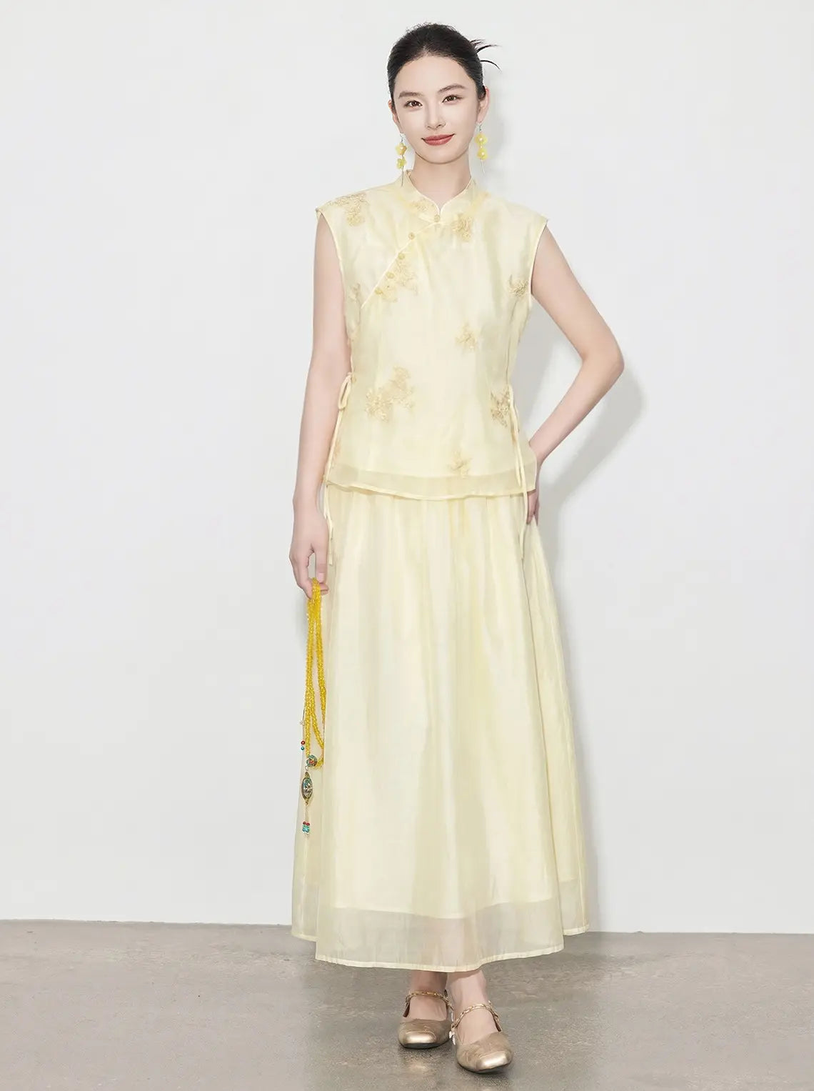 Woman wearing a light yellow sleeveless top and long skirt with floral details on a white background